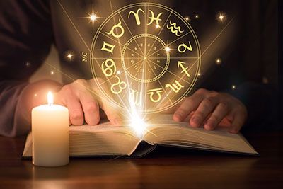 Hands placed on an open book with zodiac signs in a circle and stars  floating above.